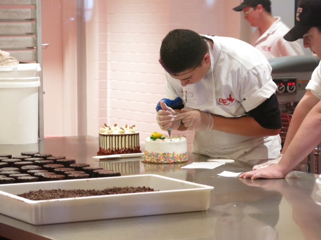 Ao fundo da cozinha, mais bolos sendo confeitados