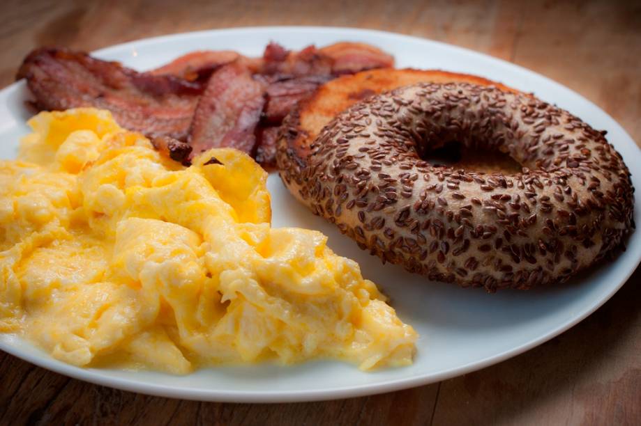 O café da manhã é composto de bagel na chapa, ovos mexidos e bacon, e vem com um potinho de cream cheese O café da manhã é composto de bagel na chapa, ovos mexidos e bacon, e vem com um potinho de cream cheese