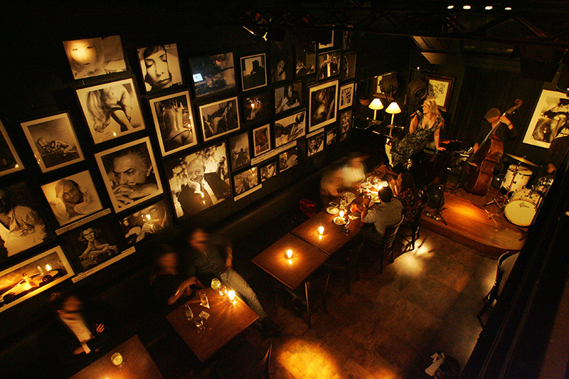 No mezanino e no salão, bem escurinhos, as mesas se voltam para o palco No mezanino e no salão, bem escurinhos, as mesas se voltam para o palco