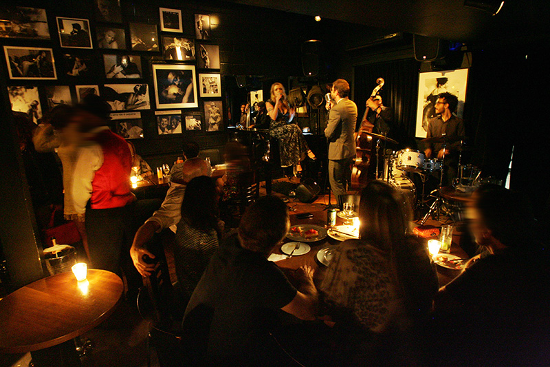 No mezanino e no salão, bem escurinhos, as mesas se voltam para o palco No mezanino e no salão, bem escurinhos, as mesas se voltam para o palco