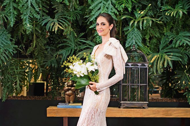 A jornalista Chris Flores posa com buquê e vestido branco. Ao fundo, plantas por toda a parede.