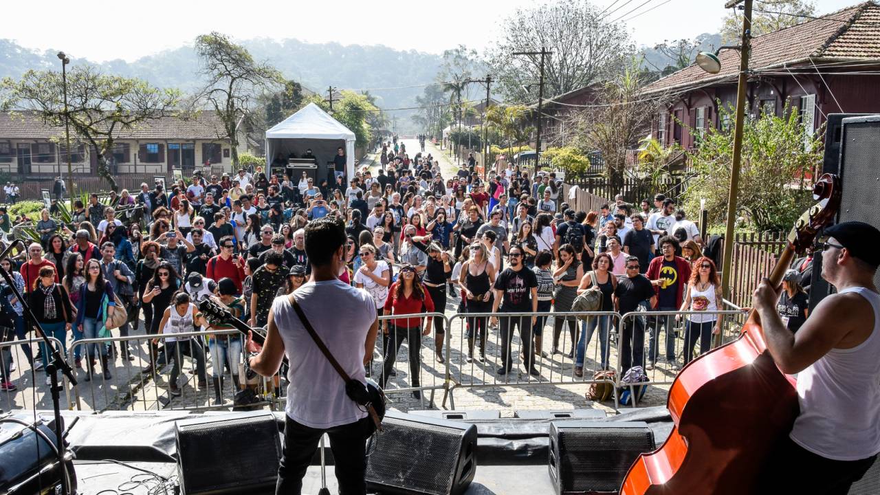 Imagem mostra palco visto de cima, com homem tocando guitarra de costas e, na sua frente, grande público em cenário montanhoso e arborizado aos fundos