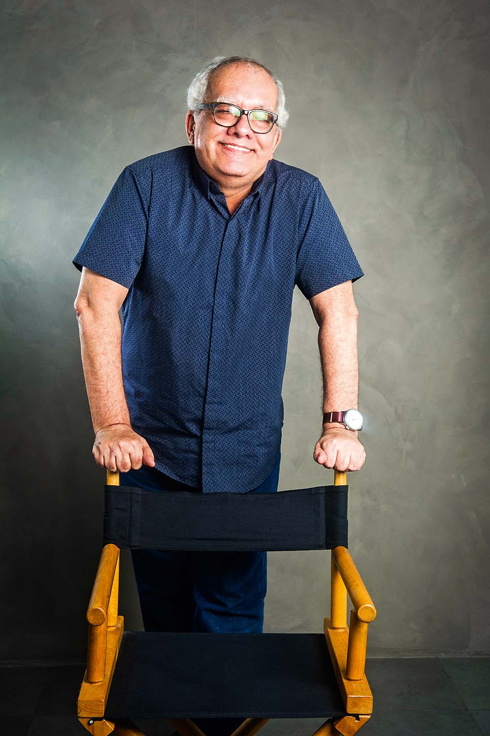 Retrato de Artur Xexéo de pé apoiado em cadeira.