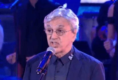 Imagem mostra Caetano Veloso de camisa escura e paletó azul, de óculos e cantando