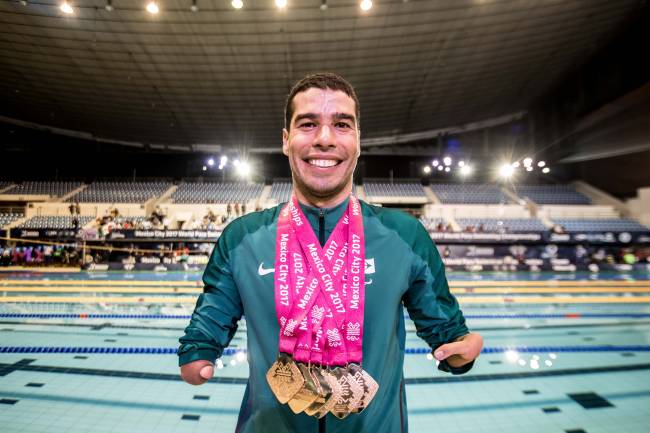 Imagem mostra Daniel Dias com diversas medalhas no pescoço. Ele está na frente de piscina olímpica