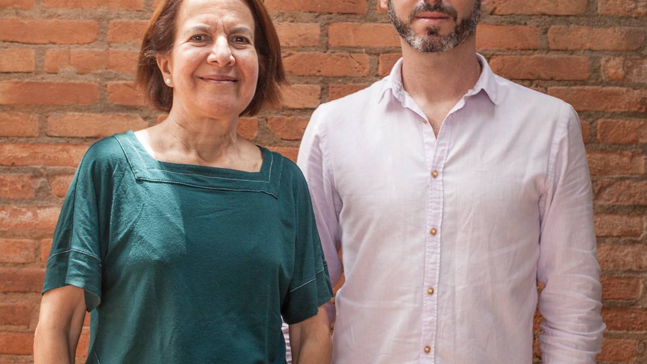 Isabel e Gonçalo posam de pé lado a lado. Ela veste camiseta verde e calça preta. Ele veste camisa rosada e calça escura. Ao fundo, tijolos aparecem na foto.