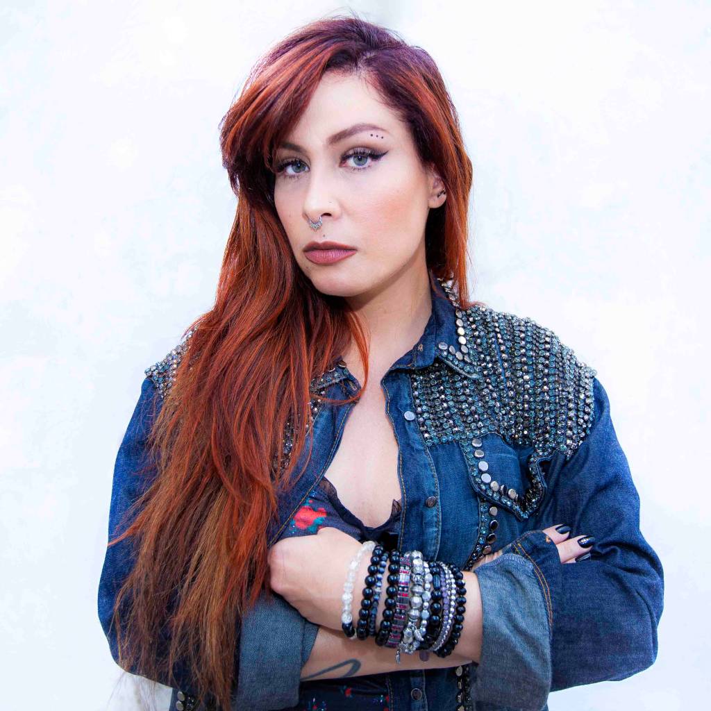 Pitty. Pitty posa de braços cruzados com jaqueta jeans e cabelo vermelho.