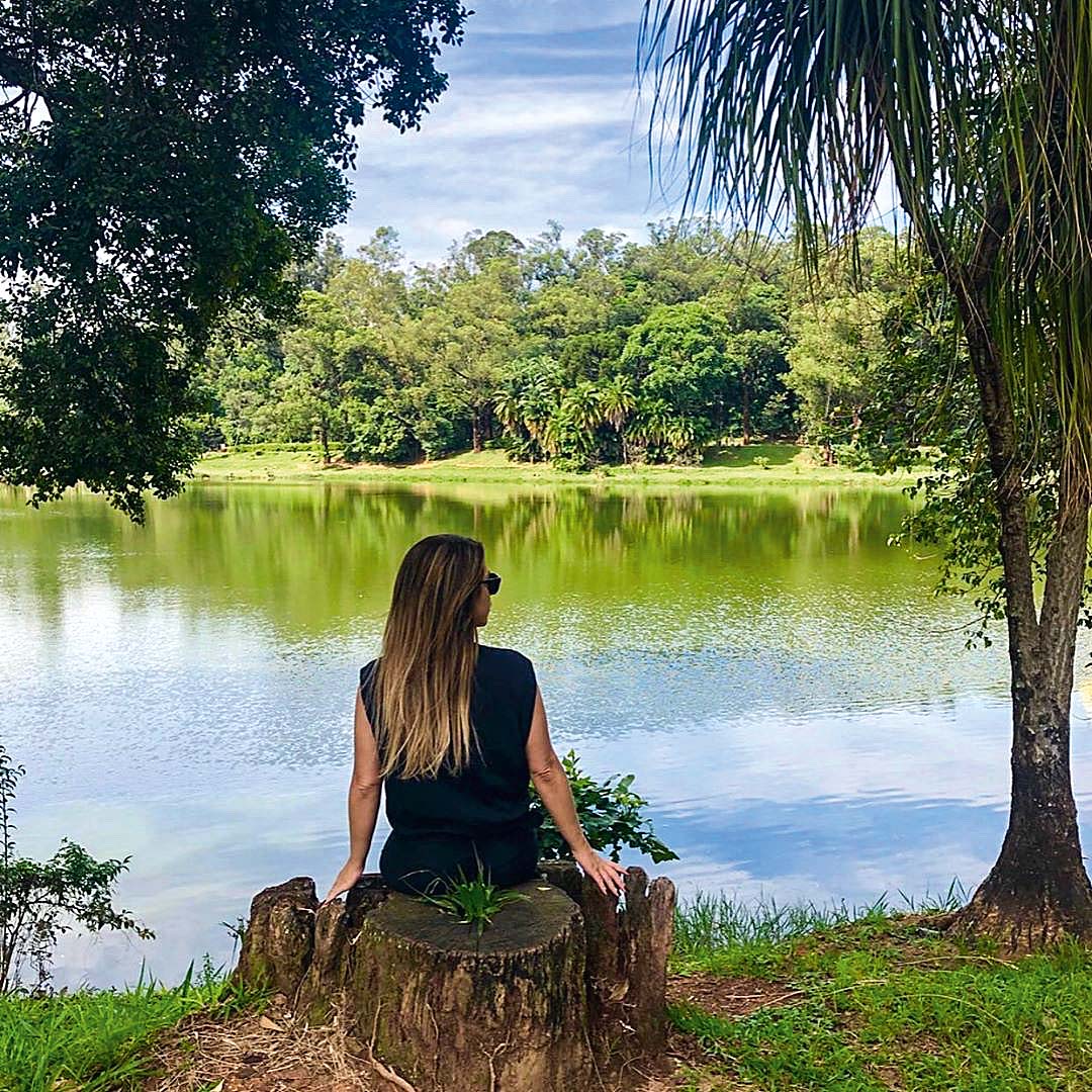 Imagem mostra um dia ensolarado. Uma mo&ccedil;a est&aacute; sentada na grama em um parque.