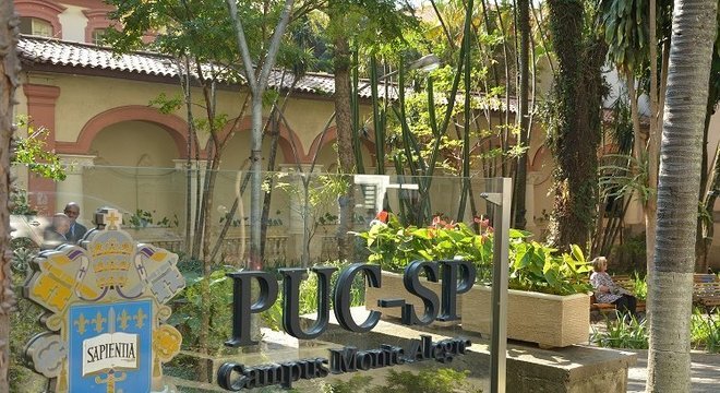 Vidraça exibe as letras PUC-SP em frente a jardim do campus.