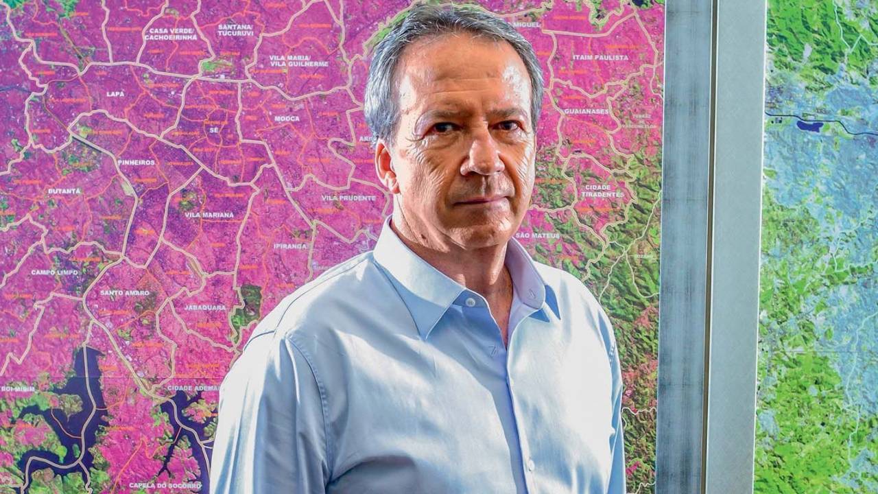 Imagem mostra Edson Aparecido olhando para a câmera, posicionado na frente de um mapa da cidade de São Paulo