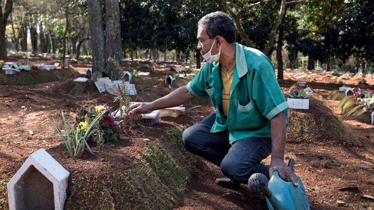 Coveiro mexe em arranjo em cova em cemitério. Ele segura um regador