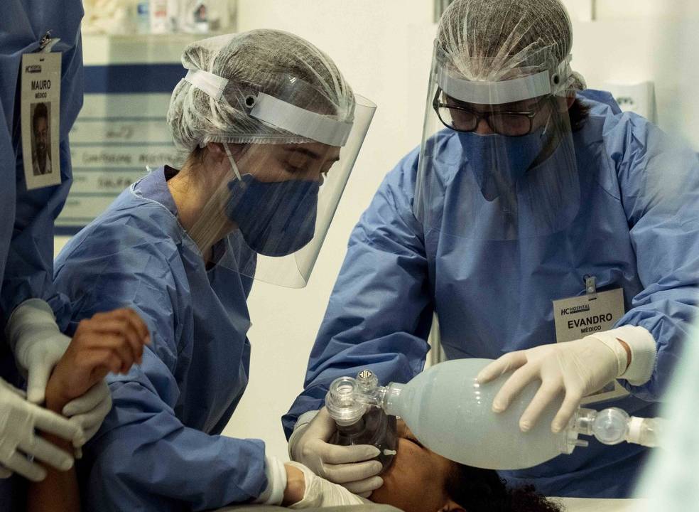 hospital de campanha durante pandemia. Na imagem, dois médicos, com equipamentos de proteção, estão em volta de uma cama com um paciente deitado. Um deles, na cabeça da cama e do paciente, segura um balão de ar.