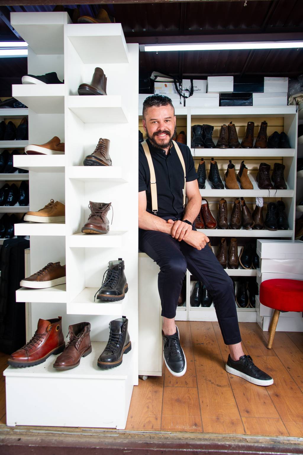Thiago Landes (Pallatta) Homem com barba posa encostado em prateleira repleta de sapatos.