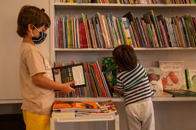 Foto mostra estante de livros infantis e duas crianças de máscara mexendo nas obras.