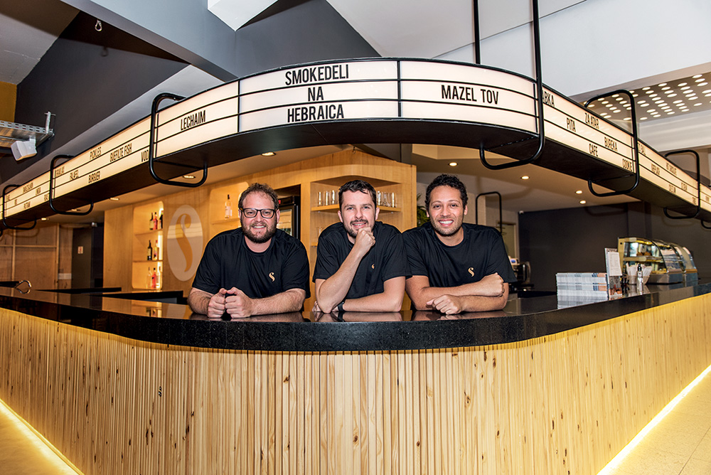 Debruçados em um balcão circular com letreiro luminoso em estilo de cinema antigo, os sócios Gabriel Baum, Bruno Wolfsdorf e Rafael Zolko olham para a câmera.