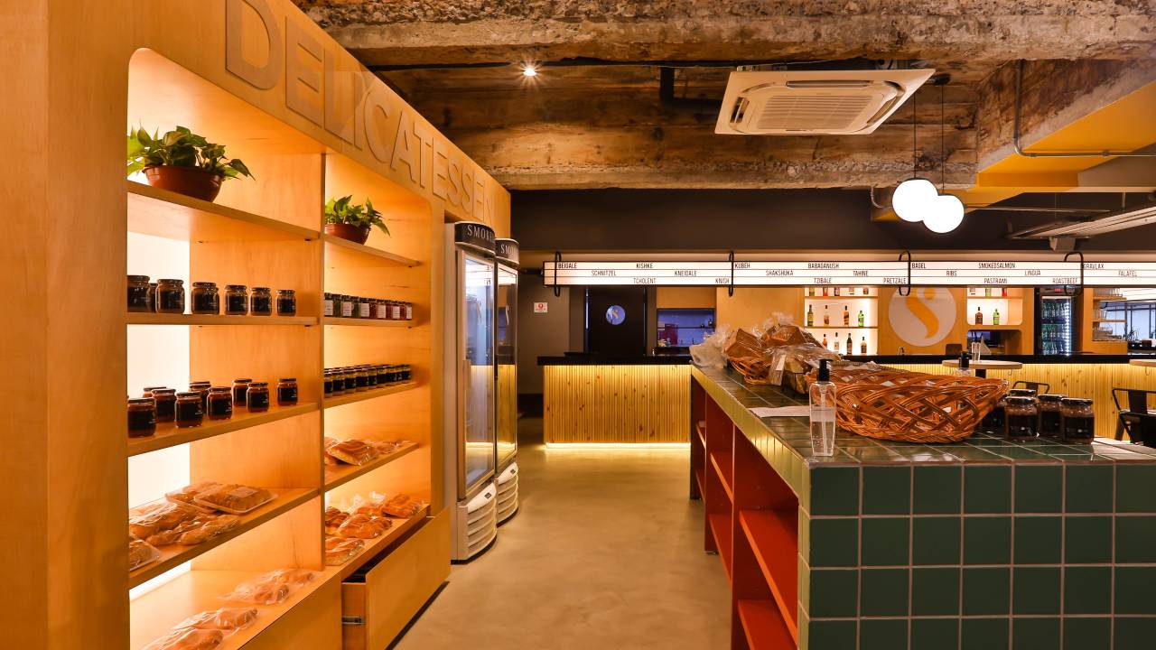 Prateleiras exibem produtos kosher iluminados e alinhados com balcão verde e vermelho à direita, em ambiente de empório da lanchonete.