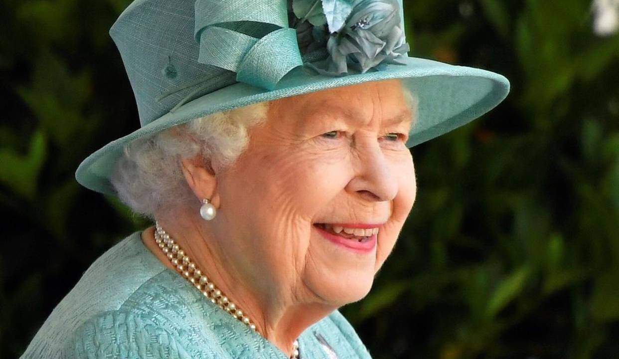 A imagem mostra Rainha Elizabeth II sorrindo. Ela usa chapéu e vestes de cor verde azulado