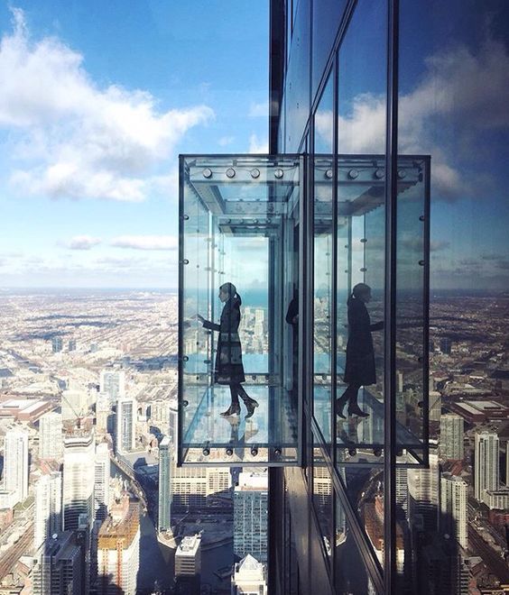 Mirante Sampa Sky – Centro (DIVULGAÇÃO) Box de vidro surge projetado para fora de um prédio com vista da cidade ao fundo. Uma mulher aparece dentro do espaço envidraçado olhando para baixo.