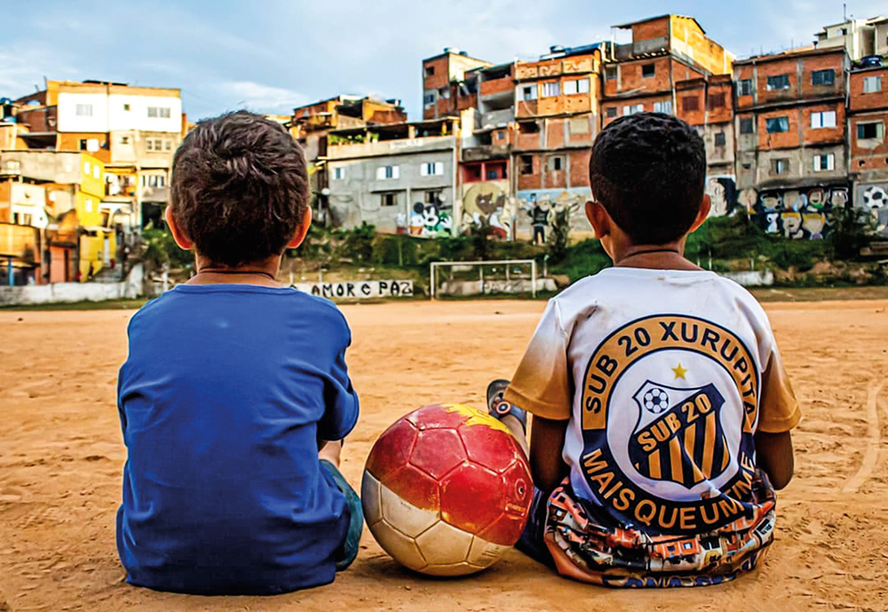 AAA 95903408_189171668791654_1624408173054630952_n.jpg (1) Duas crianças sentadas em um campo de terrão, com uma bola vermelha entre elas. A visão é das costas dela, e ao fundo elas tão observando um gol vazio