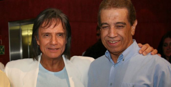 Foto mostra o cantor Roberto Carlos utilizando um paletó, abraçado com o seu irmão Lauro Braga, que usa uma camisa