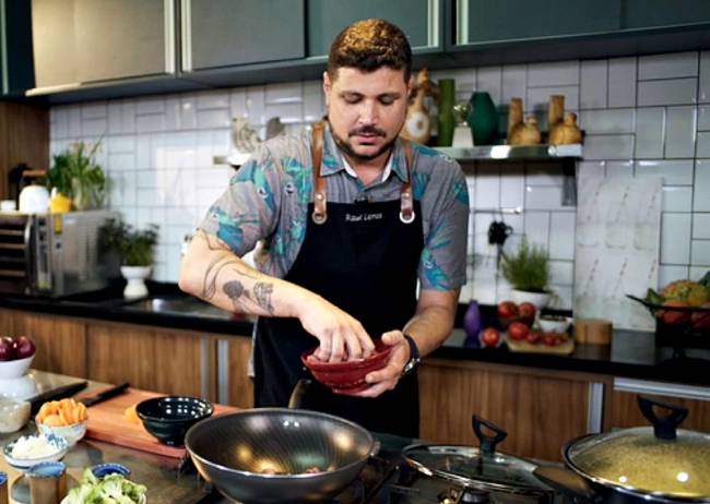 Homem com avental culinário jogando ingredientes de uma cumbuca para uma frigideira funda numa cozinha