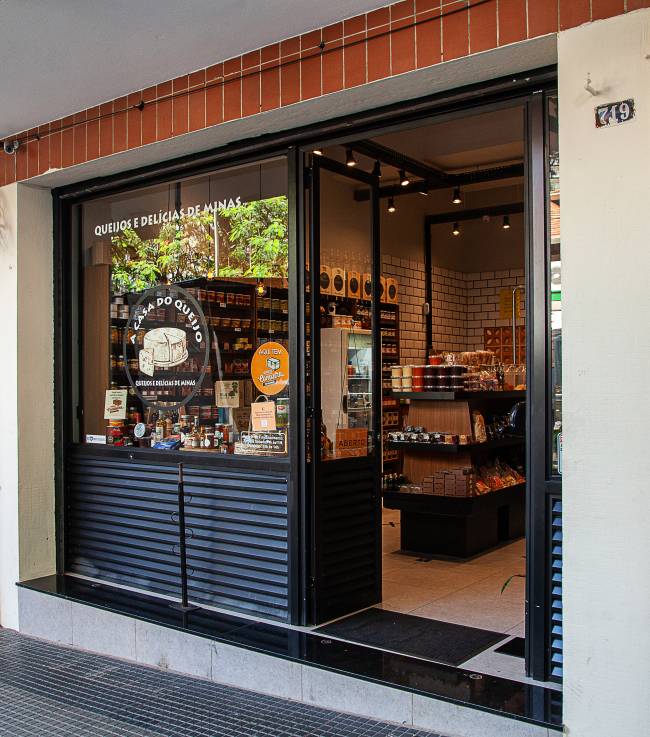 A Casa do Queijo – Achados Elo Fachada da loja A Casa do Queijo. Porta à esquerda com vitrine de vidro sobre estrutura metálica preta à esquerda.