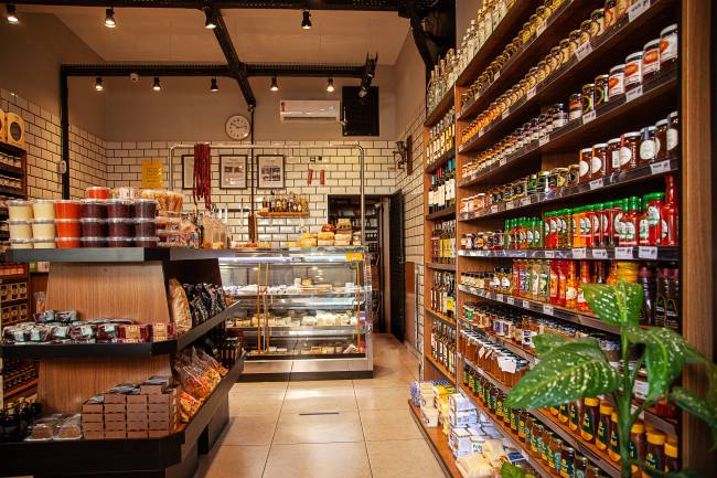 A Casa do Queijo – Achados Elo Interior da loja A Casa do Queijo. Estufa de queijos ao fundo, gôndola com pimentas e geleias à esquerda e estante de madeira com doces caipiras à esuqerda.