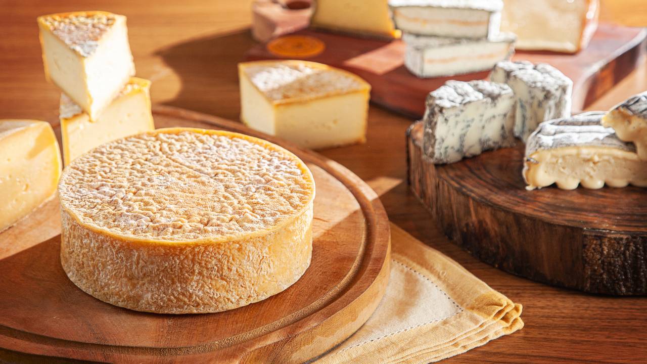Mesa de madeira com tábuas de queijo. À esquerda em primeiro plano, queijo inteiro redondo sobre tábua do mesmo formato, pedaços em quartos cortados ao fundo. À direita em segundo plano, queijos cobertos por mofo azul cortados ao meio.