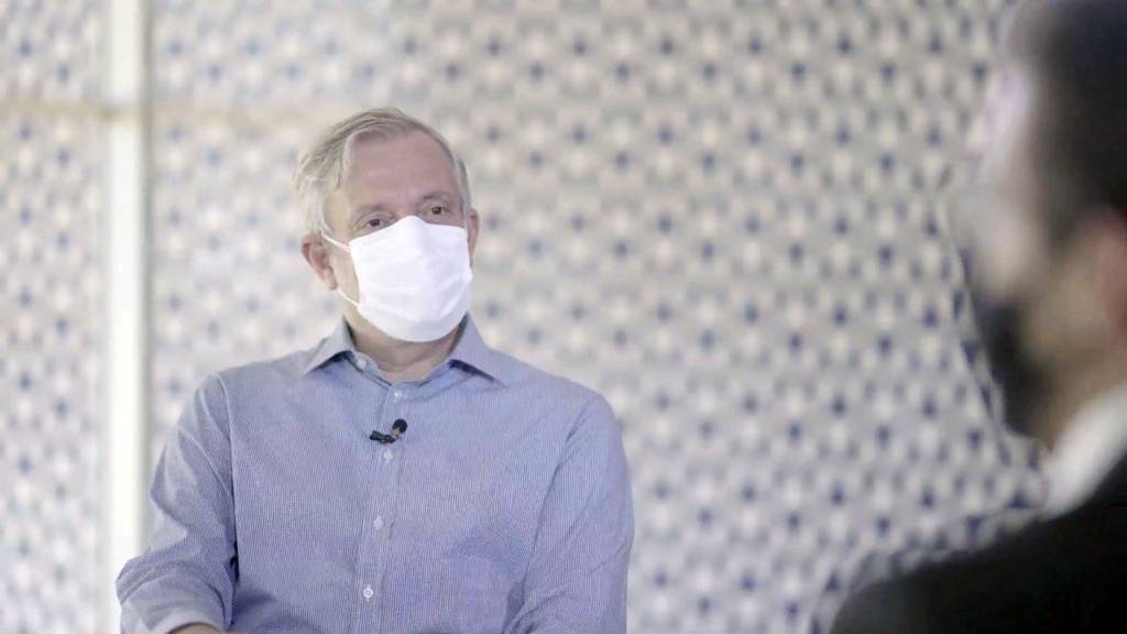Alvaro entrevista Gustavo Mendes gerente geral de medicamentos da Anvisa (Globo-Divulgação) Repórter Álvaro Pereira Jr. aparece com máscara branca e camisa azul em frente a silhueta de Gustavo Mendes, gerente-geral de medicamentos da Anvisa.