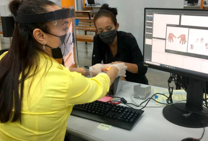 A imagem mostra atendimento em unidade do Poupatempo: mulher estende as mãos para impressão digital