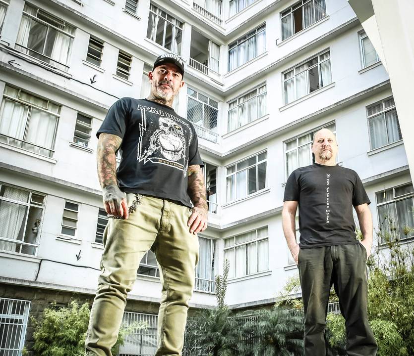 Os dois homens, de roupas casuais. A foto é vista de baixo para cima. Os dois estão de pé em cima de um banco de concreto, com um prédio ao fundo
