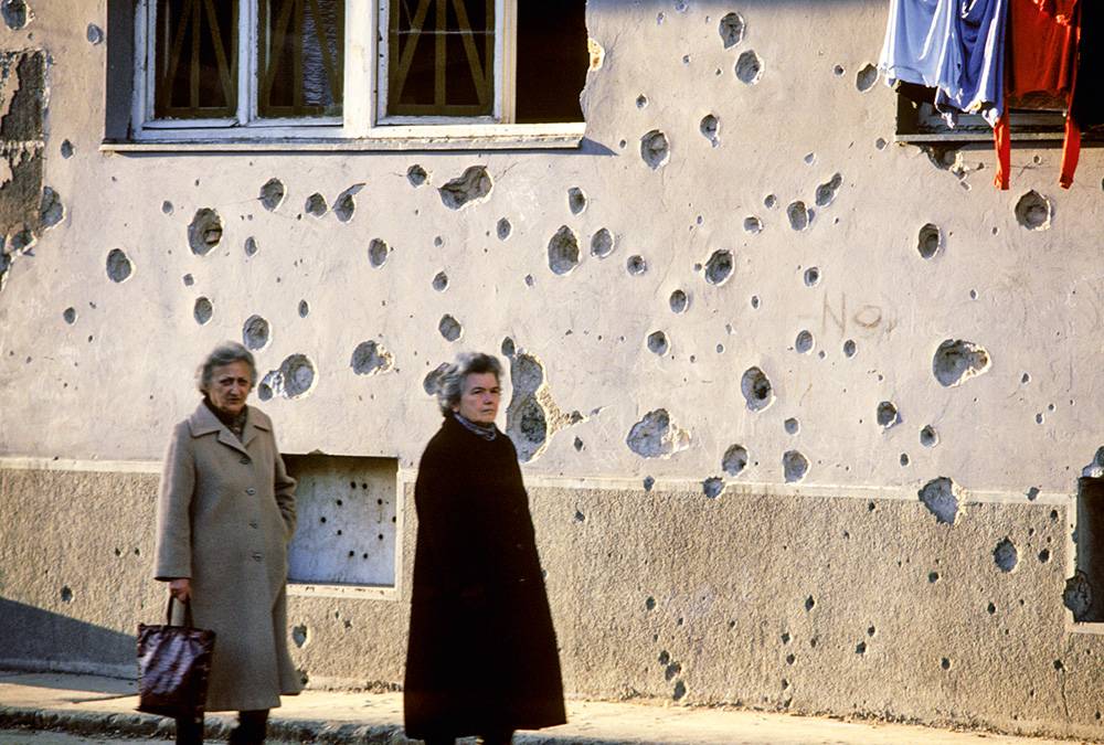 Marcas de bombardeios nas paredes e carro destru&iacute;do abandonado na rua: cidade-ru&iacute;na e ecos da viol&ecirc;ncia