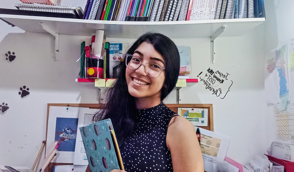 pequenos-cientistas-giovanna.jpg Giovanna posando para foto sorrindo e segurando caderno