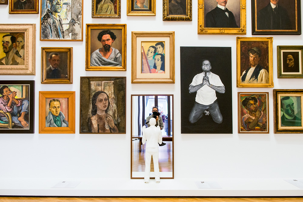 Levi Fanan_Pinacoteca.jpg Vários quadros dispostos em uma parede branca, um deles é um espelho e da para ver o fotógrafo tirando a foto