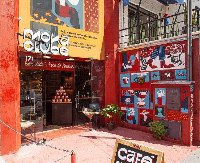Achados Elo – Moka Clube Foto angulada pelo canto esquerdo da fachada da loja Moka Clube. Paredes vermelhas cobertas por pôsteres azul e branco do lado direito e porta de vidro do centro para a esquerda.
