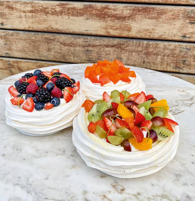 Três pavlovas postadas em cima de uma mesa de granito, uma coberta de mix de frutas, outra de frutas vermelhas e a última, mais ao fundo, de caqui.