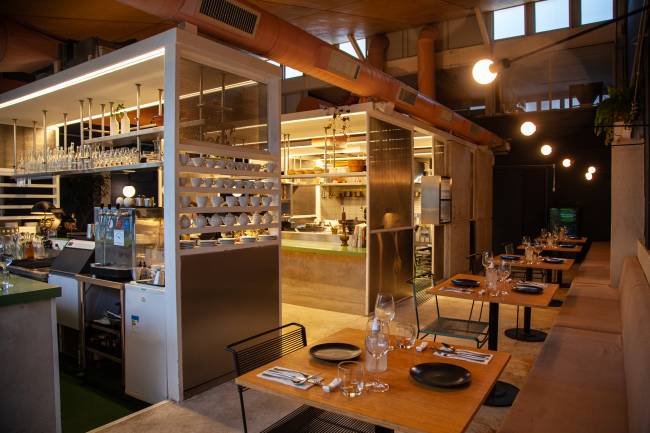Corrutela ambiente restaurante com mesas postas à direita e cozinha aberta por janela de vidro ao fundo à esquerda