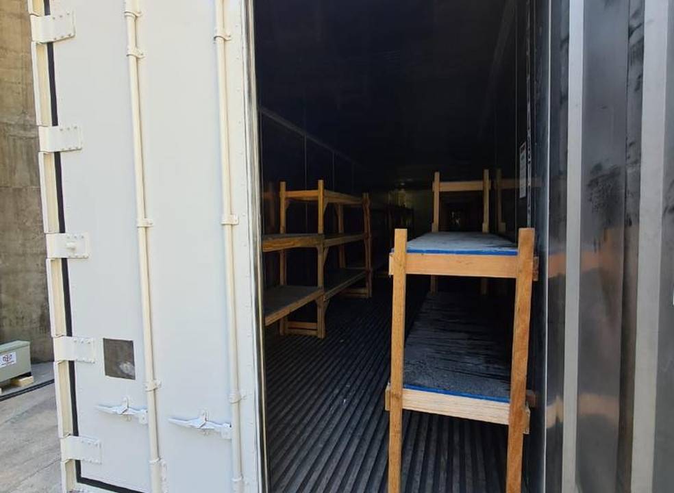 Imagem mostra contêiner com uma das portas abertas. Dentro dele, beliches para guardar os corpos