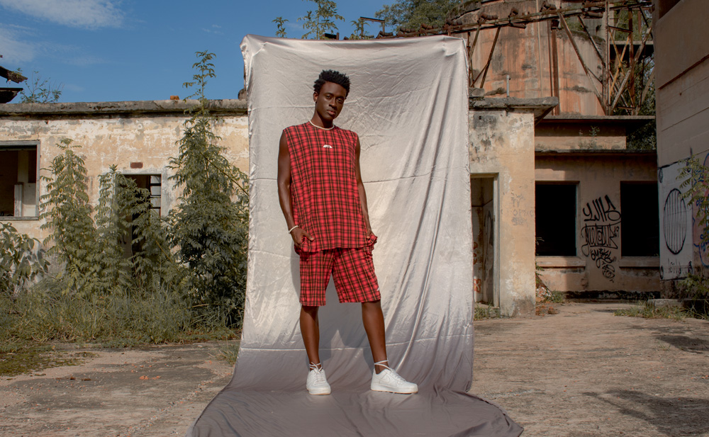 Jovem negro posa com conjunto de roupas (shorts e regata) da mesma estampa, quadriculada vermelha e preta. Ao seu fundo, um lençol branco estendido, em meio ao quintal de uma casa