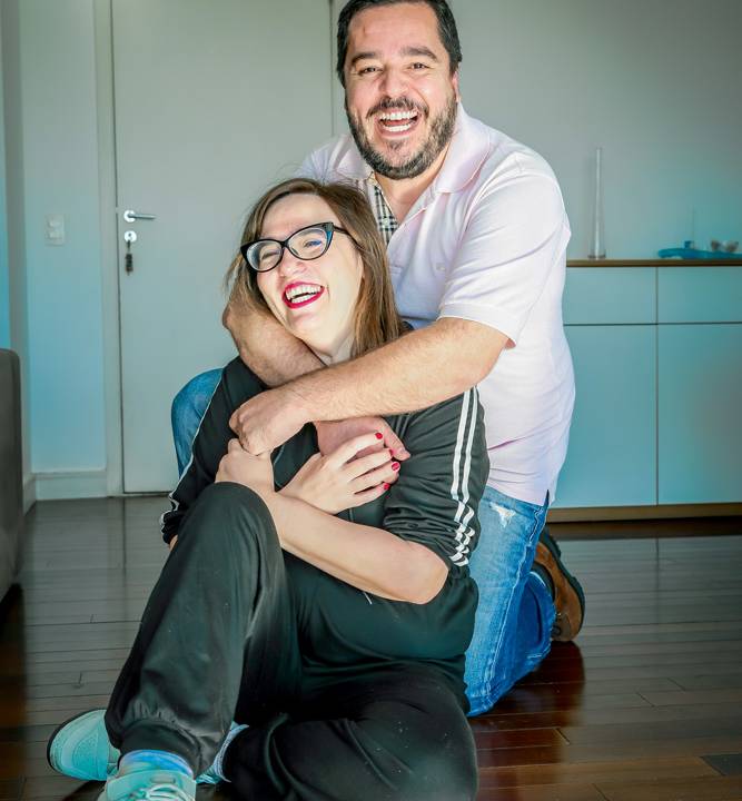 adriana e luciano no chão de sua casa, ela sentada sendo envolvida pelos braços dele, que está ajoelhado atrás dela
