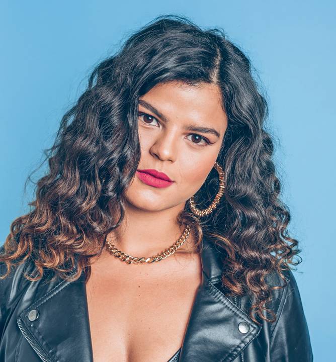 A atriz e cantora Amanda Magalhães posa em fundo azul, com cabelos cacheados, brincos de argola e batom vermelho. Veste jaqueta de couro.