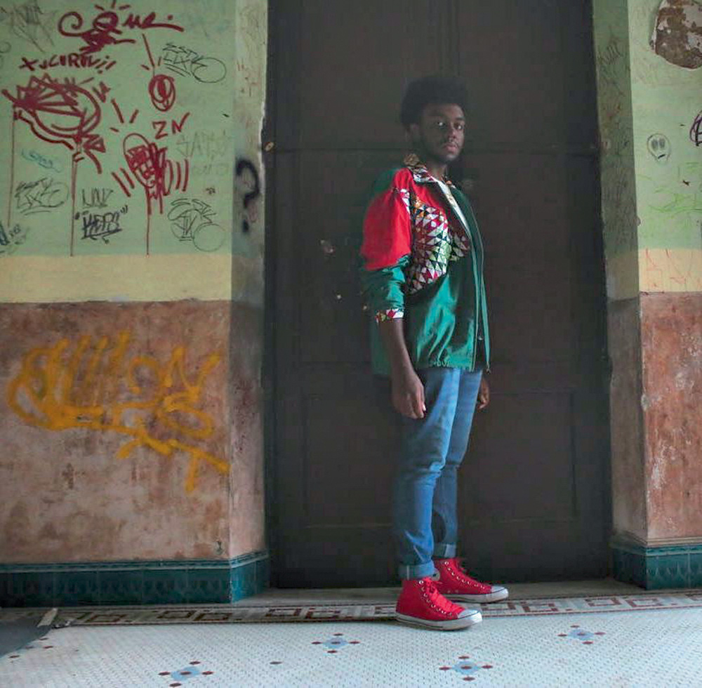 Créditos @vaicremoso.jpg Jovem negro posa em frente a uma porta, em cenário com pinturas de tinta nas paredes