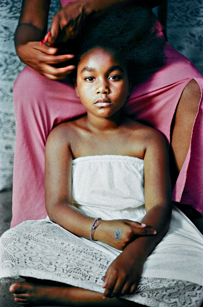 DanieleQueiroz.jpg A imagem mostra uma criança tendo os cabelos crespos penteado pela mãe. A menina está sentada no chão, apoiada na cadeira que a mãe está sentada e passando o pente.