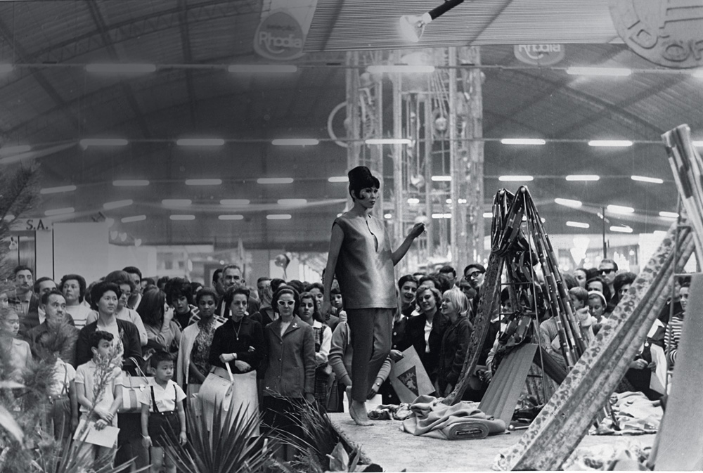 Desfile da FENIT no Pav. Ibirapuera_1.jpg A imagem em preto e branco mostra a modelo em uma passarela no meio da feira de automóveis. O local está lotado de pessoas.
