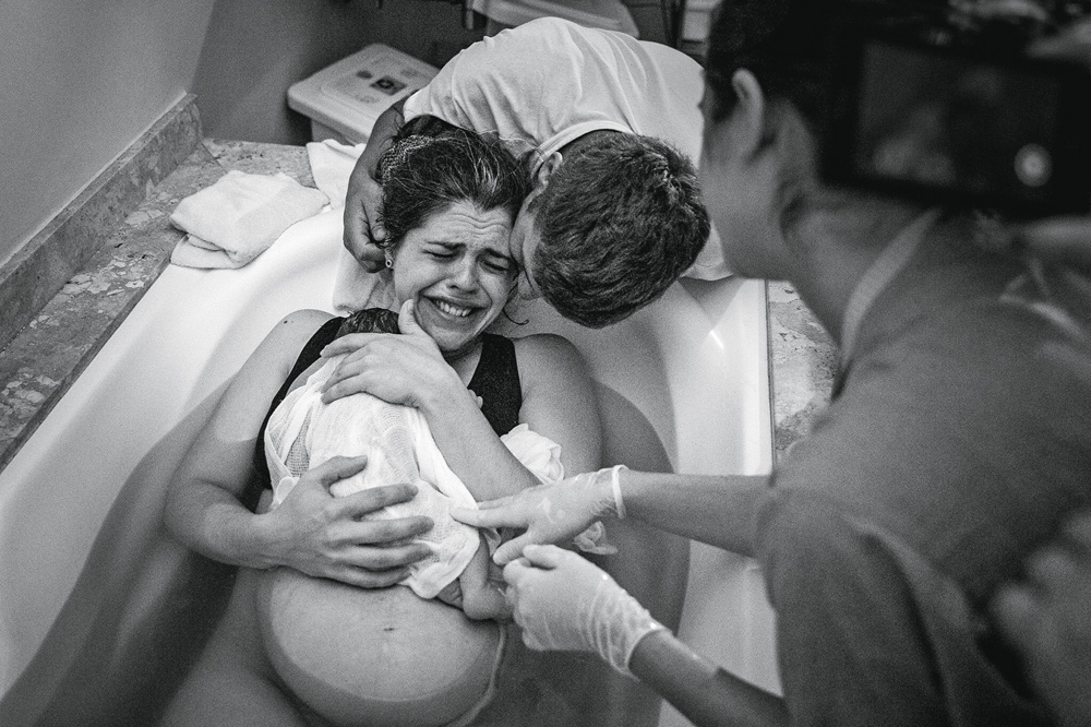 Diana Mendes Luz.jpg Foto em preto e branco mostra mulher na banheira dando a luz à bebê. Neném está em seu colo, um homem a abraça e ela chora