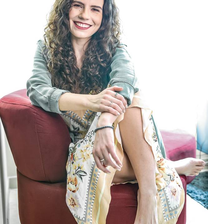 A imagem mostra Helena, sentada em uma poltrona vermelha em um fundo claro. Ela sorri para a câmera.