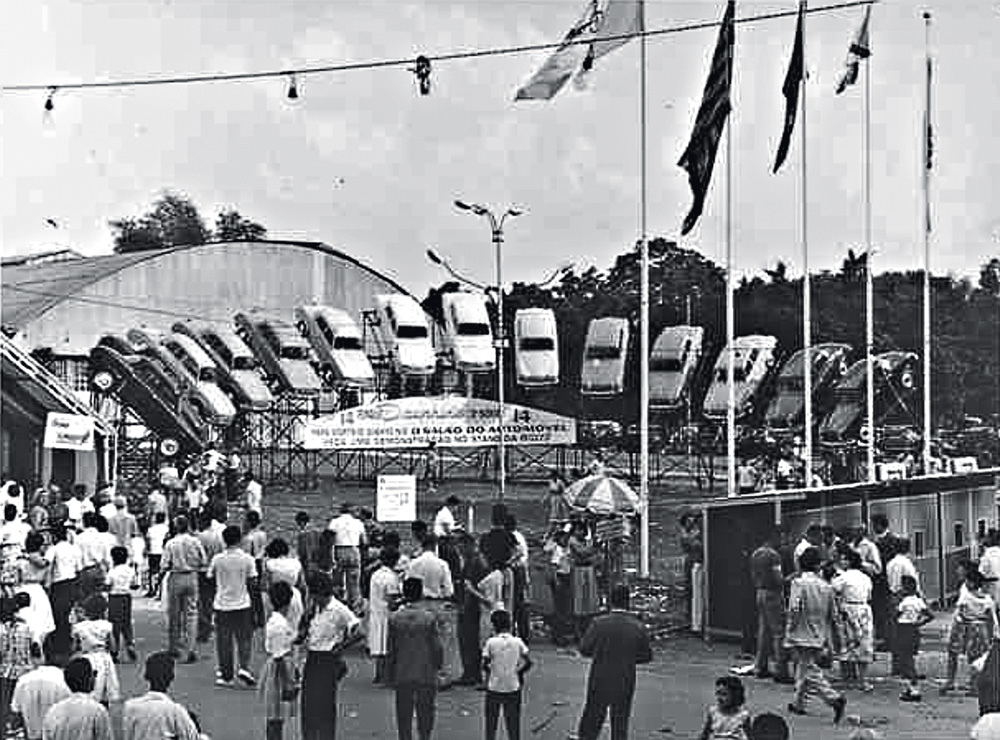 II Salão do Automóvel.jpg A imagem em preto e branco mostra a entrada do salão do automóvel. É uma fachada lotada de pessoas e, ao fundo, há carros erguidos com a frente voltada para o chão.