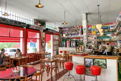 Sal&atilde;o amplo de bar com mesas e cadeiras de madeira &agrave; esquerda, ao lado de janelas, e balc&atilde;o com banquetas vermelhas ao fundo do lado direito.