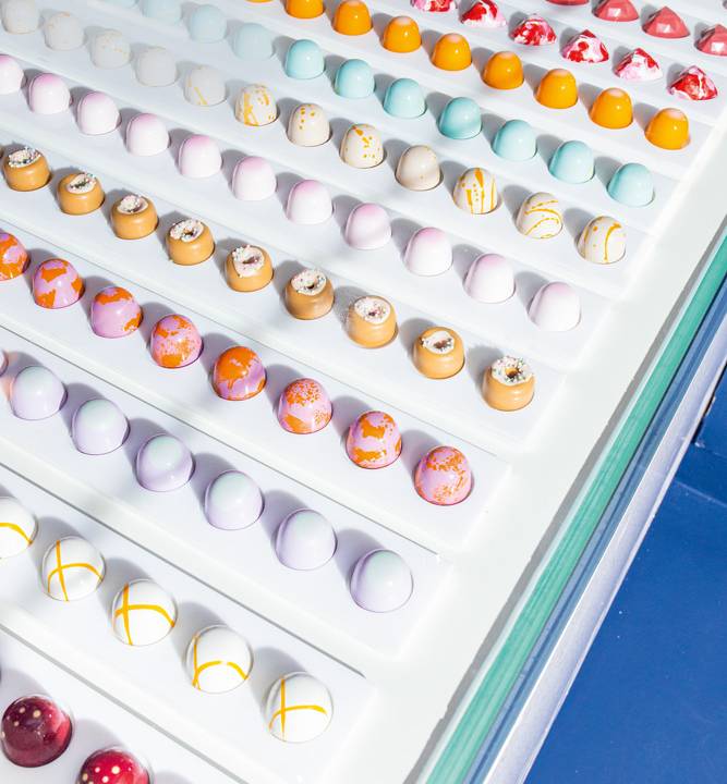 Foto de uma vitrine diagonal, com fundo branco, fileiras de bombons alinhados pintados à mão da Mica Crafted Chocoaltes.