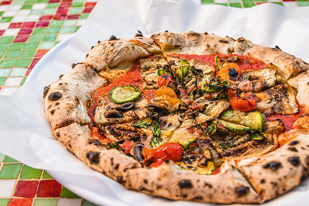 Pizza vegetariana sobre papel branco com molho de tomate, berinjela, abobrinha, cogumelo, cebola-roxa e manjericão.